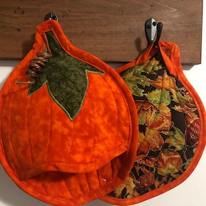 May include: Two quilted pumpkin-shaped pot holders. One is solid orange with a green leaf and stem detail. The other has a fall leaf pattern on a dark background. Both are hanging on hooks.