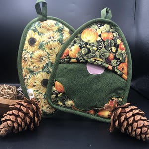 May include: Two quilted pot holders. One features sunflowers on a yellow background, the other has a green gnome design with a pumpkin and floral pattern. Both have green trim and hanging loops.