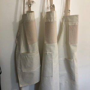 May include: Three striped aprons hanging on a wooden rack. The aprons have a cream background with thin gray stripes, a large front pocket, and a tan patch on the chest. The straps and ties are a light beige color.