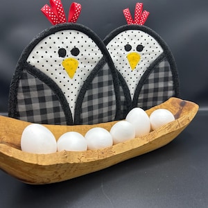 May include: Two chicken-shaped pot holders with black and white gingham and polka dot patterns. They have yellow beaks and red polka dot ribbon accents. A wooden trough holds six white eggs.