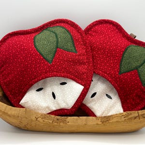 May include: Two quilted fabric pot holders shaped like apple slices. Each features a red dotted top section, a white dotted bottom section representing the apple flesh, and two green fabric leaves stitched onto the top red portion.