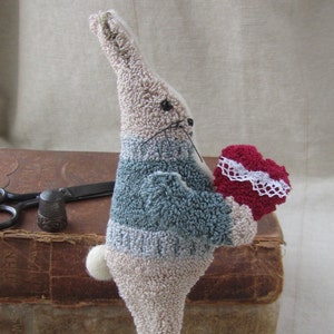 May include: A handmade plush bunny figurine in beige, blue, and red, holding a red heart with white lace trim. The bunny has black whiskers and is attached to a copper-colored base with a blue ribbon. The figurine is placed in front of a stack of old books.