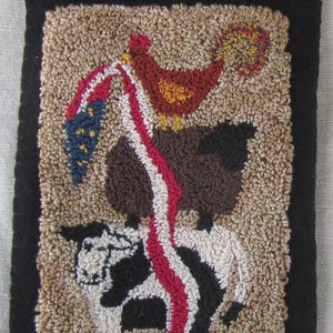 Op de afbeelding: Handgehaakte vloerkleed met een koe, schaap en kip, met een vlagachtig ontwerp. Het vloerkleed heeft een zwarte rand en een neutrale achtergrond. De koe is zwart en wit, het schaap is bruin en de kip is rood.