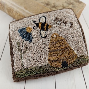 May include: Square, textured rug with a bee, beehive, and flower design. The rug features a bee in flight, a brown beehive, and a blue flower. The text "1847" is above the beehive. The rug has a brown border and a green base.