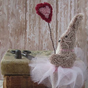 May include: A handcrafted bunny doll wearing a pink tutu and holding a heart-shaped balloon. The bunny is light brown with pink details, and the tutu is made of white tulle. The heart balloon is red and pink. The doll is placed on old books.