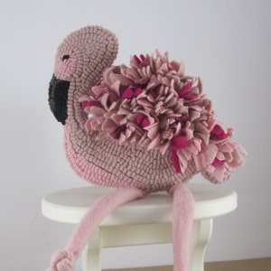 May include: A handmade pink flamingo plush toy with a textured body, black beak, and a cluster of pink and fuchsia fabric feathers. The flamingo has long, pink legs and is perched on a small white stool. A whimsical decorative item.