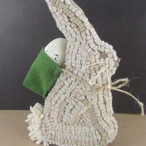 May include: A decorative Easter bunny figurine made of textured, off-white fabric. The bunny holds a speckled egg in a green pocket and has a fluffy white tail. A twine bow adds a rustic touch to this Easter-themed decoration.