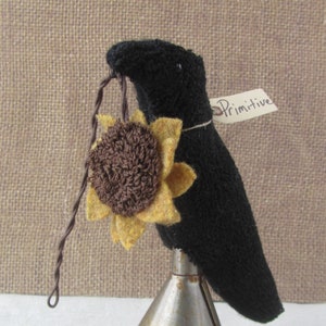 May include: A black fabric crow ornament holding a felt sunflower with brown and yellow petals. The crow is perched on a metal stand. A tag that says "Primitive" is attached to the crow.
