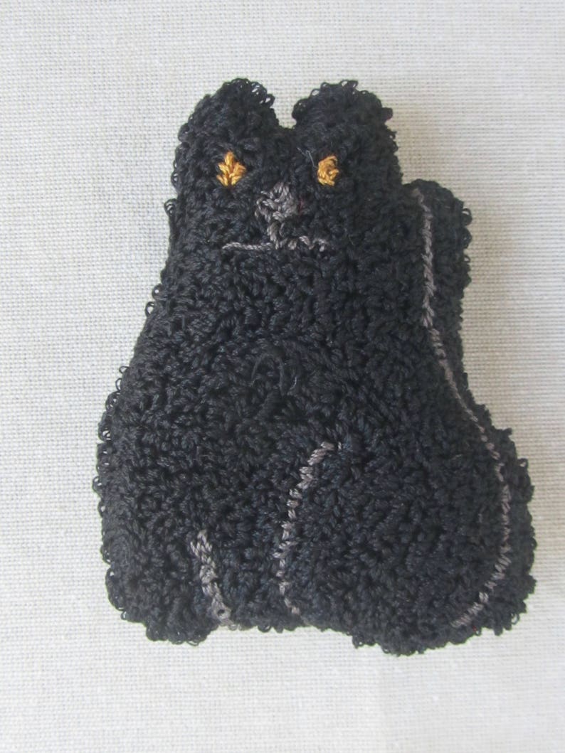 May include: A black cat-shaped textile item with a looped texture. The cat features golden-yellow eyes and a grey outline. This handmade item is likely a decorative piece or a small toy, with a whimsical design.