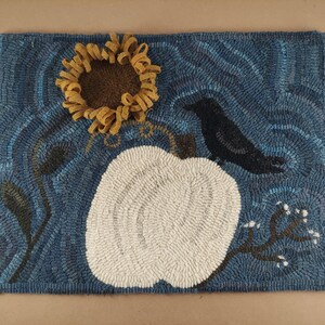 May include: A rectangular rug with a blue background featuring a white pumpkin, a black crow, and a yellow sunflower. The rug has a textured, handmade appearance, with the design elements stitched into the fabric. The overall theme is autumn or fall.