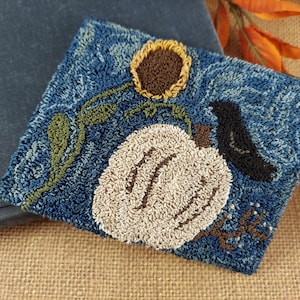 May include: A square, textured, blue textile art piece featuring a white pumpkin with brown accents, a sunflower, and a black bird. Green leaves and brown stems are also present. The artwork is placed on a burlap surface.