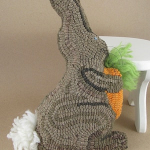 May include: A handmade, textured bunny rabbit figurine in shades of gray, holding an orange carrot with green leafy tops. The bunny has a fluffy white tail and black accents. The figurine is likely made of fabric or yarn.