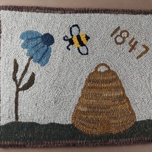 May include: Hand-hooked rug featuring a blue flower, a bee, and a beehive. The rug has the number "1847" and a brown border. The background is a light color, and the design elements are in various colors.