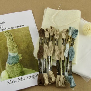May include: A punch needle pattern kit featuring a bunny design. The kit includes a pattern booklet, embroidery floss in various shades of brown, beige, and blue, fabric, and a small pom-pom. The booklet reads "Doodle Dog Designs Punch Needle Pattern Mrs. McGregor."