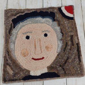 May include: A square, hand-hooked rug featuring a portrait of a person with a light skin tone, gray hair, and a dark blue hat. The rug has a brown border and a red, white, and blue design in the upper right corner. The face has a pinkish hue.