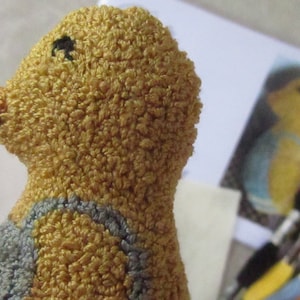 May include: A handmade, textured yellow duckling with a small black eye and a light brown beak. The duckling is wearing a blue and gray outfit. A picture of a similar duckling is in the background.