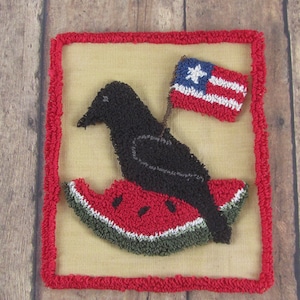 May include: A handcrafted textile artwork depicting a black crow on a watermelon slice, holding a small flag with a white star and red and white stripes. The piece is framed with a red border and set against a tan background.
