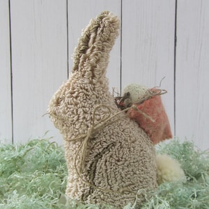 May include: A handmade Easter bunny figurine with a textured, beige body and long ears. It has a small, pink sack tied to its back with twine and a fluffy white tail. The bunny is set in green decorative grass.