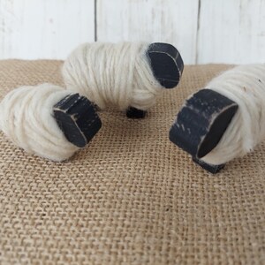 May include: Three handcrafted sheep figurines. Each sheep has a white wool body and a black wooden head and legs. The sheep are arranged on a burlap surface, creating a rustic and charming aesthetic. These decorative items are perfect for a farmhouse or country-themed decor.