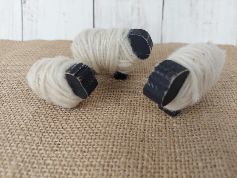 May include: Three handcrafted sheep figurines. Each sheep has a white wool body and a black wooden head and legs. The sheep are arranged on a burlap surface, creating a rustic and charming aesthetic. These decorative items are perfect for a farmhouse or country-themed decor.