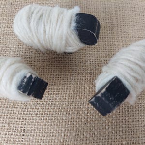 May include: Three small, handmade sheep figurines. Each sheep has a white, woolly body and a black, rectangular head. The figurines are arranged on a textured, tan surface.