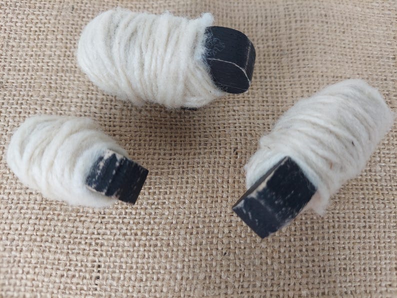 May include: Three small, handmade sheep figurines. Each sheep has a white, woolly body and a black, rectangular head. The figurines are arranged on a textured, tan surface.