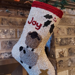 Puede incluir: Un calcetín navideño hecho a mano con fondo blanco y puño rojo. La palabra "Joy" está cosida en rojo. El calcetín presenta un diseño de oveja con detalles en negro y marrón y una estrella dorada.