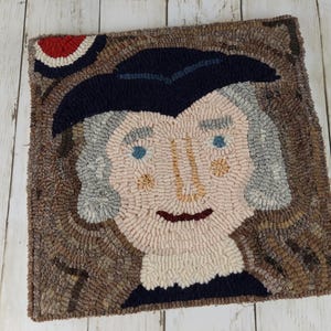 May include: Hand-hooked rug featuring a portrait of a historical figure. The rug has a square shape and depicts a face with light skin, blue eyes, and gray hair. The figure wears a dark blue hat and coat. The rug's border is brown, and the background is a light beige.