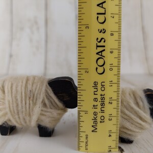 May include: Two small wooden sheep figurines. The sheep have black heads and legs, with cream-colored yarn wrapped around their bodies. A yellow ruler is placed next to the sheep, with the text "Make it a rule to insist on" visible.