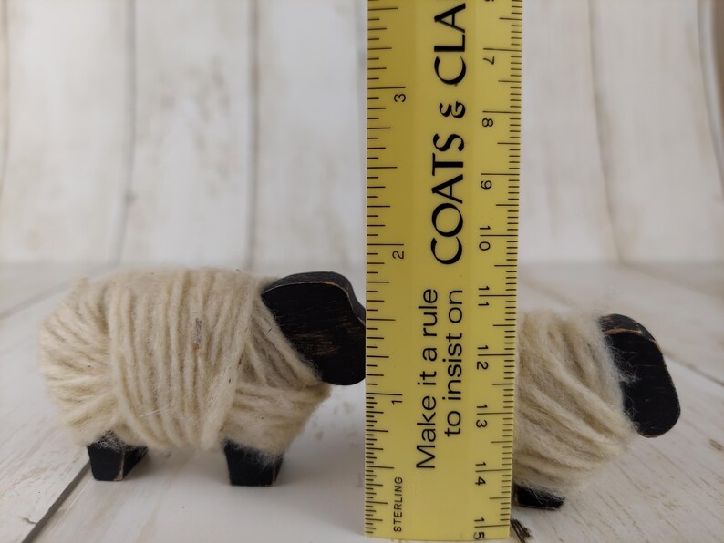 May include: Two small wooden sheep figurines. The sheep have black heads and legs, with cream-colored yarn wrapped around their bodies. A yellow ruler is placed next to the sheep, with the text "Make it a rule to insist on" visible.