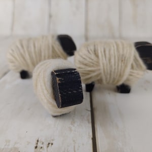 May include: Three wooden sheep figurines. The sheep have black heads and legs, with bodies wrapped in cream-colored yarn. The figurines are arranged on a white wooden surface.
