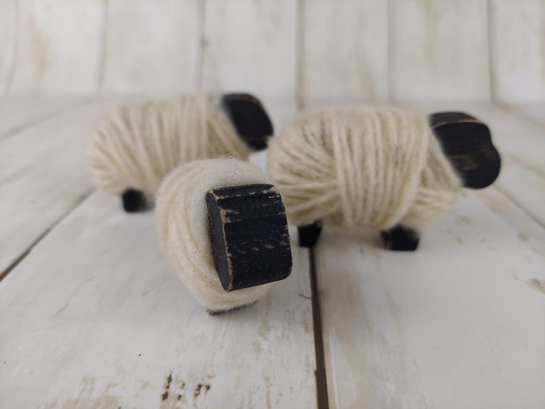 May include: Three wooden sheep figurines. The sheep have black heads and legs, with bodies wrapped in cream-colored yarn. The figurines are arranged on a white wooden surface.