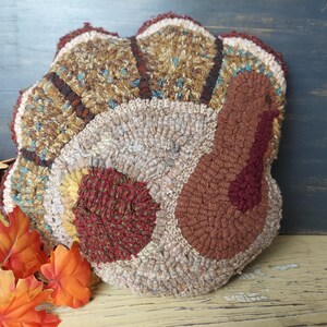 May include: Hand-hooked turkey-shaped pillow with a textured design. The pillow features a tan body, brown and burgundy accents, and a fan-shaped tail with shades of brown, tan, and teal. Autumn leaves are in the foreground.