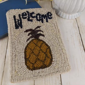 Può includere: Un tappeto rettangolare beige con un disegno di ananas e la parola "welcome" in blu navy. L'ananas è marrone con una corona marrone scuro. Il tappeto è realizzato in un materiale tessuto e si trova su una superficie di legno bianco.