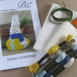 May include: A craft kit for punch needle embroidery featuring an Easter bunny design. The kit includes a pattern, embroidery floss in various colors, fabric, and other supplies. The pattern is titled "Easter Greetings."