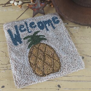 Può includere: Un tappetino di benvenuto quadrato beige con una superficie strutturata. La parola "Welcome" è in blu, sopra un disegno dettagliato di ananas nei toni del marrone e del verde. Una stella rustica e un cappello sono sullo sfondo.