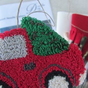 May include: A handmade red truck ornament with a green tree on top, crafted with punch needle embroidery. The truck features gray and black accents. Embroidery floss and a pattern are visible in the background, suggesting a DIY craft project.