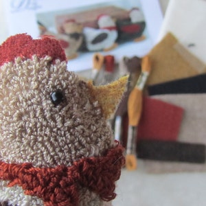 May include: A handmade chicken doll with a red comb and scarf. The chicken is made of tan and brown yarn. Craft supplies, including felt pieces and tools, are in the background. A pattern for similar chickens is also visible.