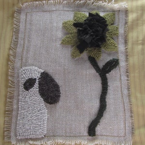 Puede incluir: Una obra de arte textil artesanal que presenta una oveja y una flor. La oveja tiene un cuerpo blanco y una oreja y un ojo negros. La flor tiene pétalos verdes y un centro oscuro. La obra de arte está sobre un fondo tipo arpillera con un borde deshilachado.