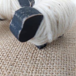 May include: Close-up of two decorative sheep figurines. The sheep have white wool bodies and black painted heads and legs. The figurines are set on a textured, tan surface.