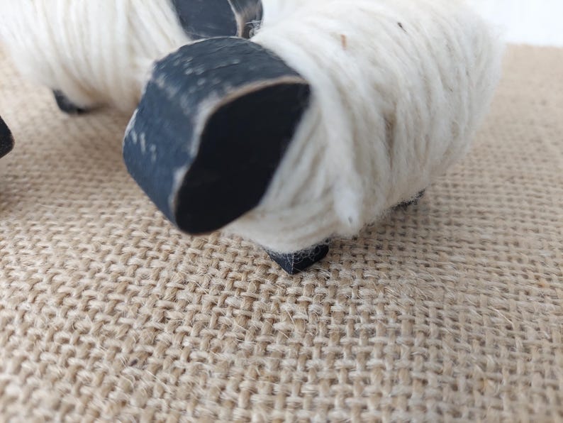 May include: Close-up of two decorative sheep figurines. The sheep have white wool bodies and black painted heads and legs. The figurines are set on a textured, tan surface.