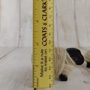 May include: Wooden sheep figurine, approximately 3.5 inches tall, with a black head and wrapped in beige yarn. A yellow ruler with black markings and text is next to the sheep. The text on the ruler says "COATS & CLARK".