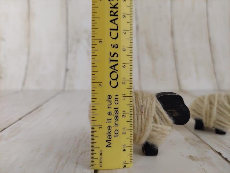 May include: Wooden sheep figurine, approximately 3.5 inches tall, with a black head and wrapped in beige yarn. A yellow ruler with black markings and text is next to the sheep. The text on the ruler says "COATS & CLARK".