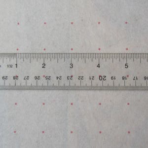 May include: A clear plastic ruler with inch markings, measuring up to 6 inches, lies over a white fabric dotted with small pink circles. The ruler is marked with numbers and fractions for precise measurements.
