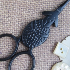 May include: Black sewing scissors with a bird-like design, featuring a textured body and a pointed beak. The scissors are next to several white buttons with two and four holes. The background is a textured, tan fabric.
