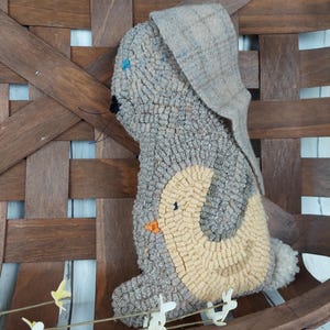 May include: A handmade, gray and tan bunny-shaped pillow with a tweed ear and a light tan belly. The bunny has a small black nose and blue eyes. The pillow is placed in a wooden basket.