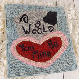 May include: Hand-hooked wool wall hanging with a light blue background. The design features a grey sheep, the word "Woolo", and a red heart with the words "You Be Mine". The heart is outlined in pink.