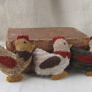 Op de afbeelding: Drie handgemaakte stoffen haanbeeldjes in bruin-, beige- en roodtinten. Elke haan heeft een rode kam en lellen, met een getextureerd, gelust stoffen lichaam. Ze zijn gerangschikt voor een vintage boek.
