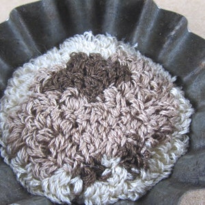 May include: A handmade, crocheted item in a fluted metal tin. The crocheted piece features a textured design with shades of cream, tan, and brown. The metal tin has a scalloped edge. The item appears to be a decorative or functional piece.