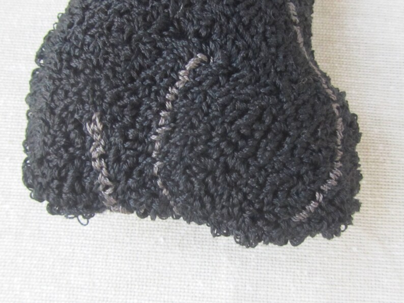 May include: Close-up of a black crocheted item with a textured surface. The item has a rounded shape and is edged with a lighter grey stitching, creating a subtle contrast. The crochet work appears dense and detailed.